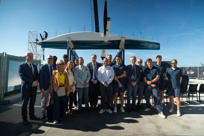 Foto di gruppo di autorit&agrave;, ospiti e membri del team davanti alla barca a foil La Roche-Posay sospesa in cantiere.