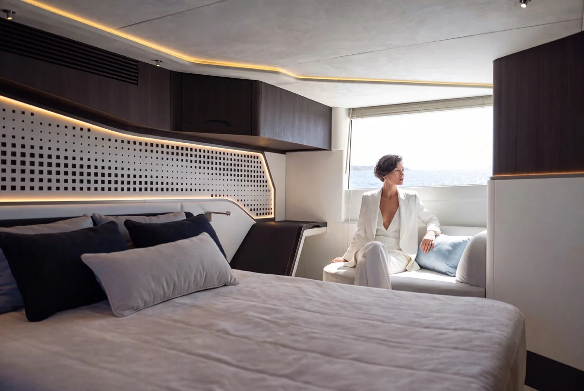 Interno cabina di yacht di lusso con letto e donna seduta vicino al finestrino, atmosfera elegante e design moderno.
