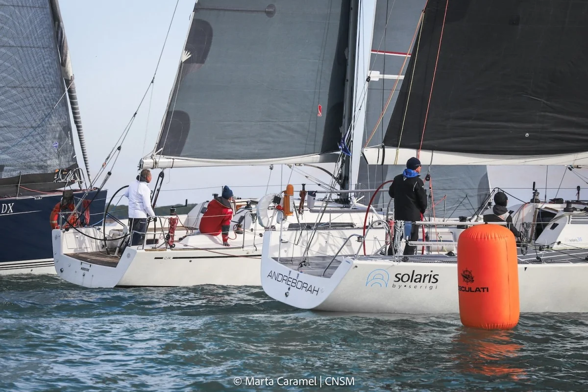 Equipaggi a bordo di barche a vela molto vicine alla boa arancione, con poppa di Andreborah e Solaris; presente il credito fotografico.