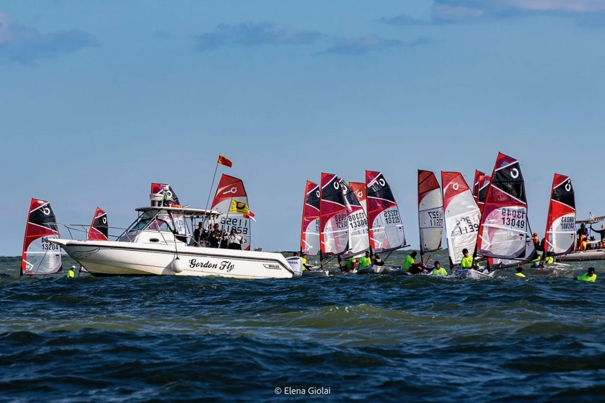 Imbarcazione d'appoggio 'Gordon Fly' affiancata da numerosi piccoli scafi con vele rosse durante la regata.