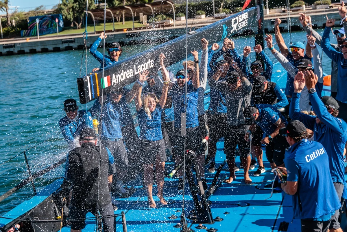 Celebrazione a bordo con spruzzi d'acqua, bottiglie e squadra esultante sul ponte della barca.