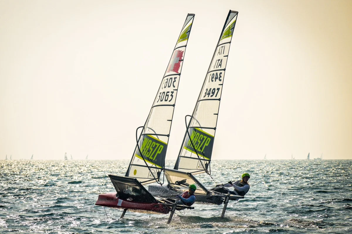 Due derive foiling in volo sull'acqua in controluce, con vele numerate e velisti con caschi verdi; atmosfera dorata.