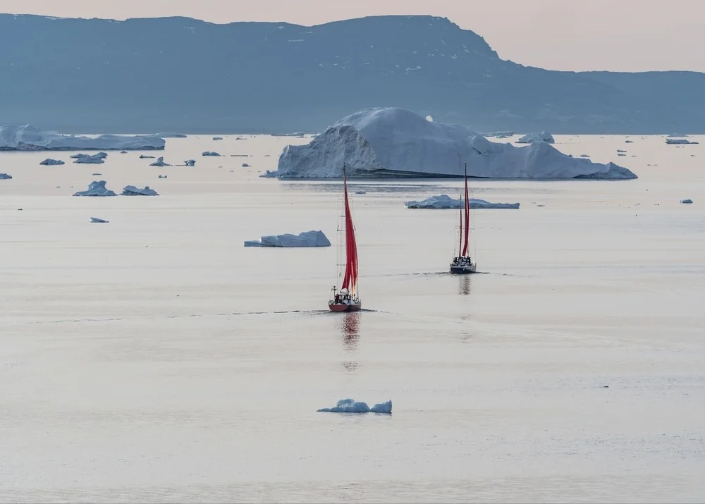Due barche a vela con vele rosse che navigano tra iceberg in mare calmo, composizione panoramica nautica.