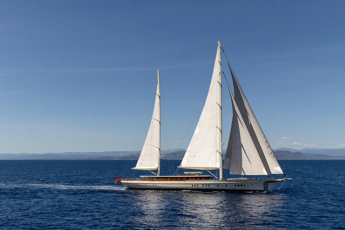 Ketch a vela bianco in navigazione sotto cielo blu, vista laterale a mare aperto. Orizzontale, pulita, ma barca piccola nel fotogramma.
