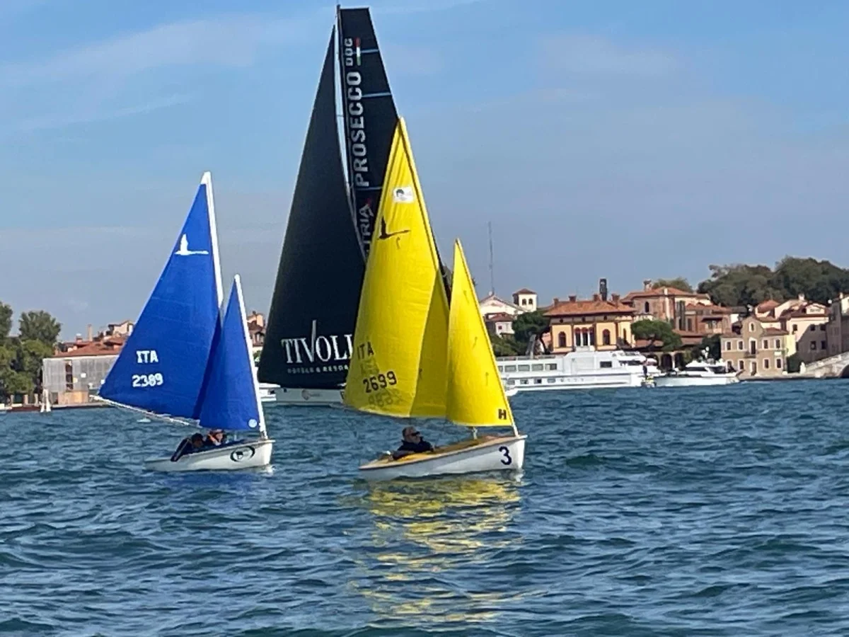 Barche a vela blu e gialle (Hansa) davanti a un grande maxi yacht nero con scritta Prosecco, sullo sfondo case veneziane. Formato 4:3.