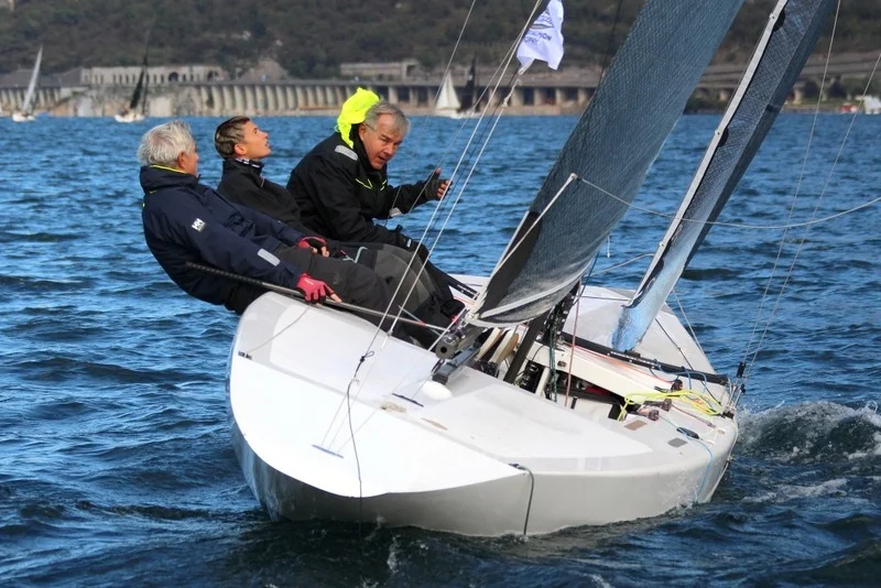 Tre velisti sbandati a prua di una deriva/chiglia moderna in navigazione sull'acqua mossa, con vela scura.