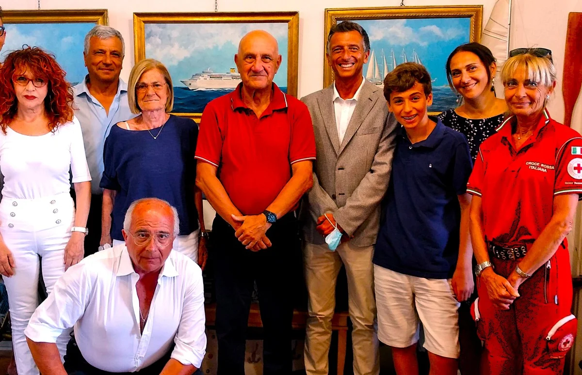 Foto di gruppo in interni con più persone (cerimonia o incontro), quadri marini sullo sfondo e presenza di soccorritori.
