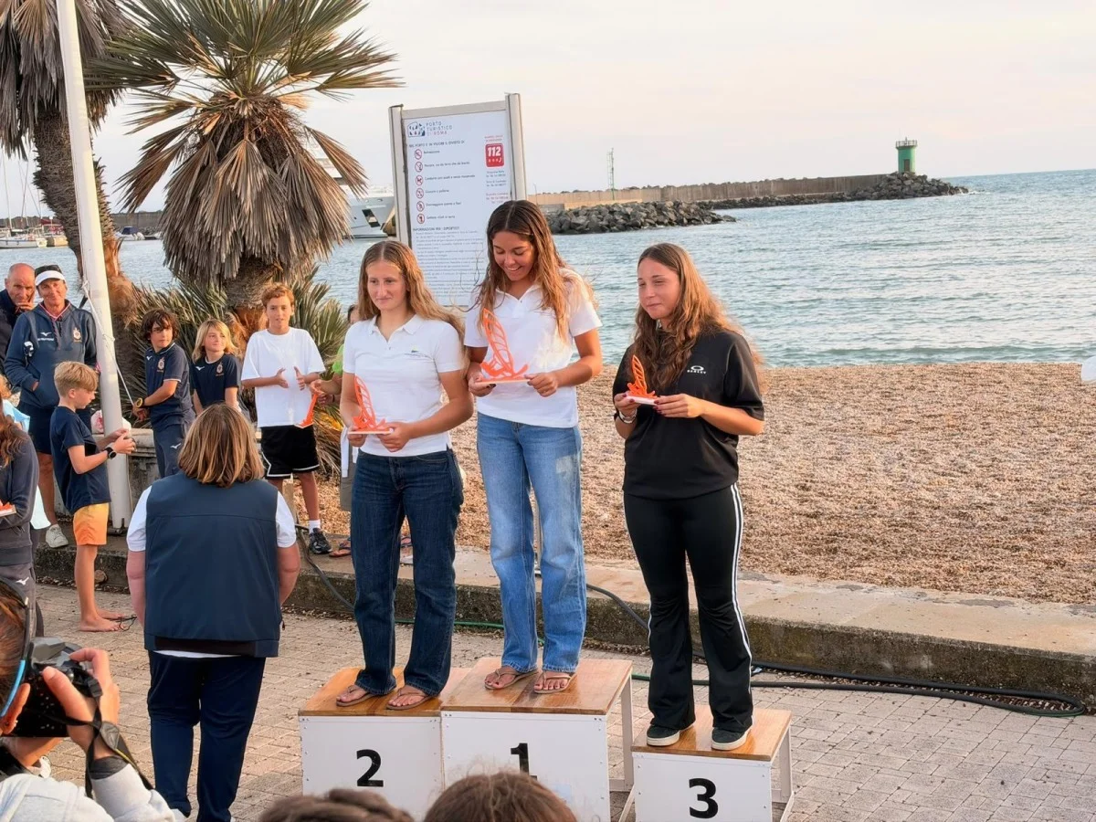 Premiazione sul podio con tre ragazze che ricevono i trofei, sullo sfondo spiaggia e molo.
