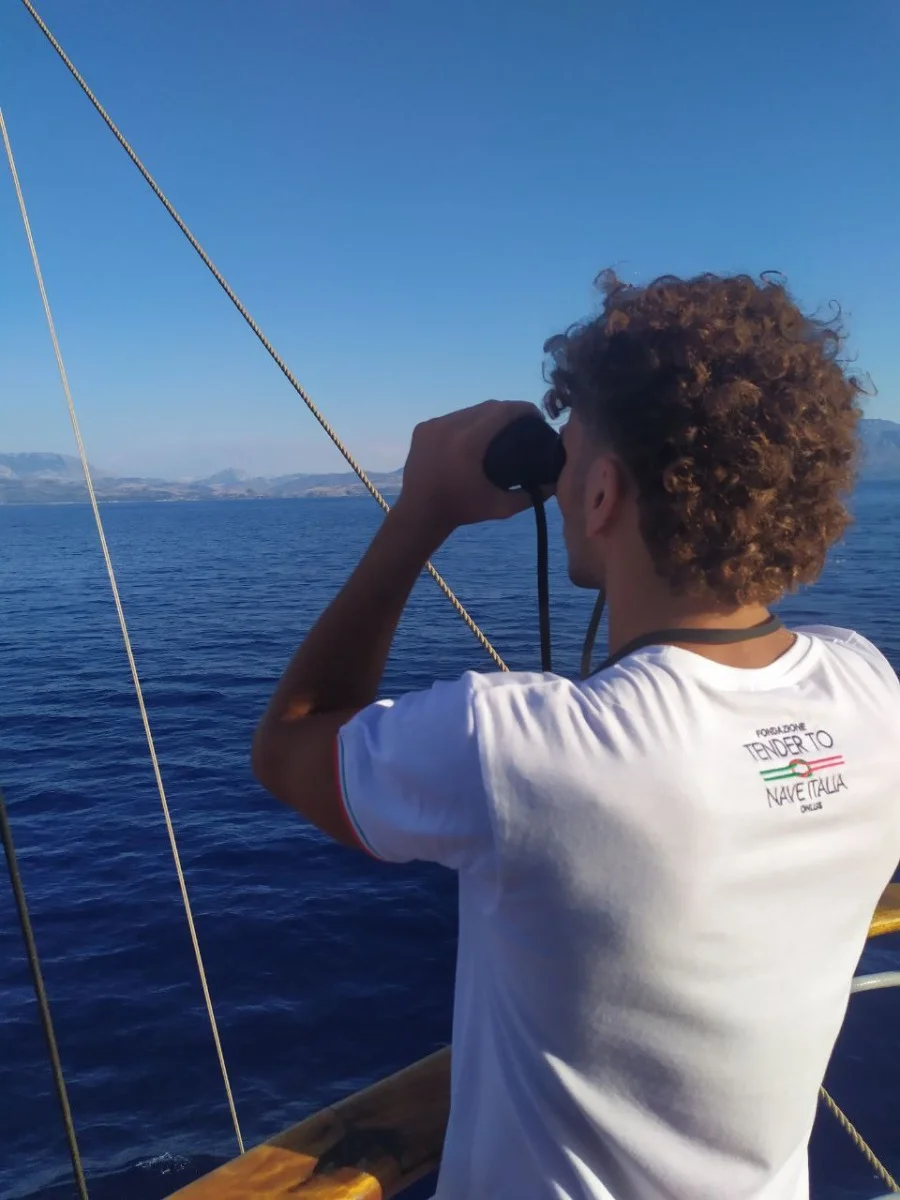 Persona di spalle con binocolo a bordo, guarda il mare aperto; foto verticale che mostra attività di bordo e abbigliamento dell'equipaggio.