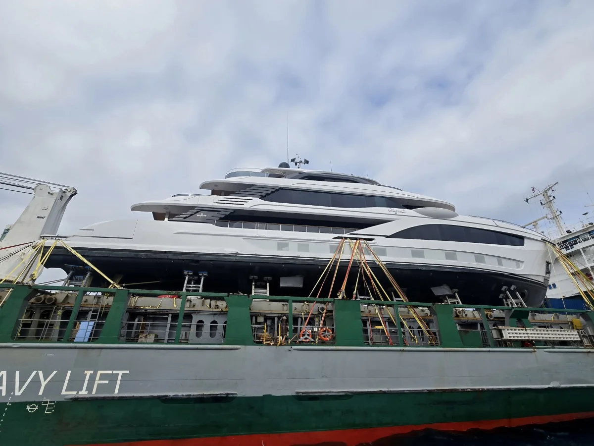 Yacht bianco caricato sul ponte della nave Heavy Lift, assicurato con cinghie gialle; cielo nuvoloso, inquadratura dal basso.