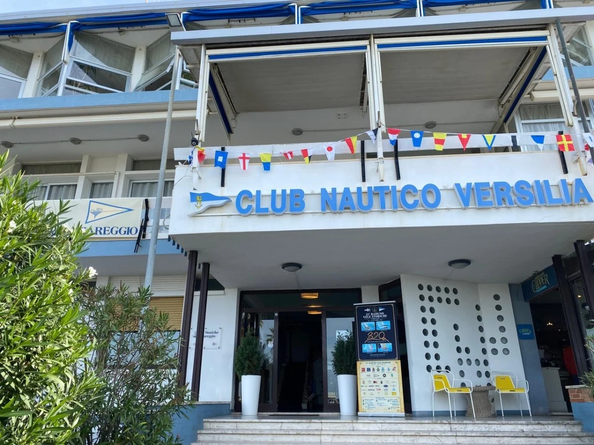 Ingresso del Club Nautico Versilia a Viareggio, con insegna blu, bandierine nautiche del codice segnali, piante e sedie gialle davanti all'entrata.