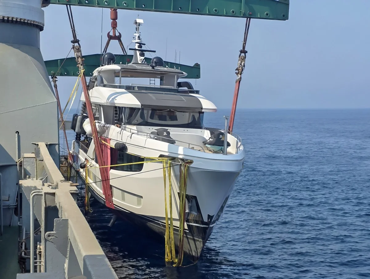 Yacht bianco sollevato da una gru con fasce gialle e rosse durante il varo dalla nave cargo, sullo sfondo del mare aperto.