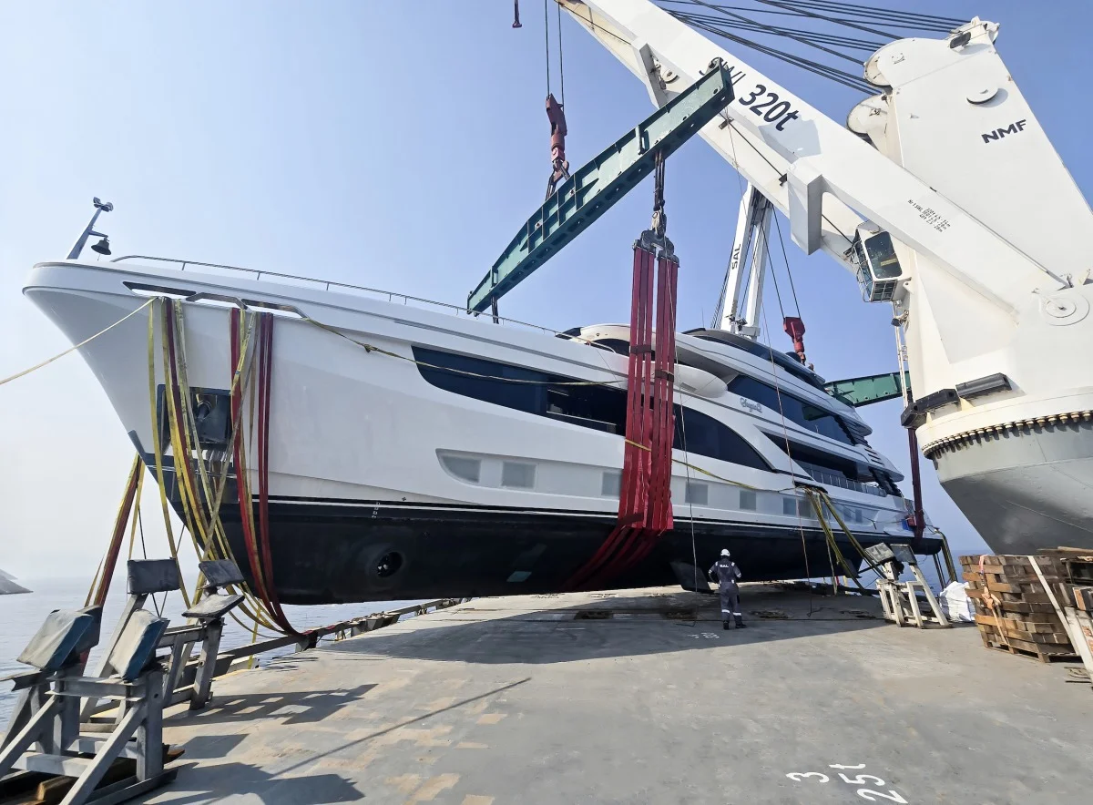Yacht sospeso dalla gru da 320 t sul ponte della nave da trasporto, con fasce rosse e un operatore in basso.