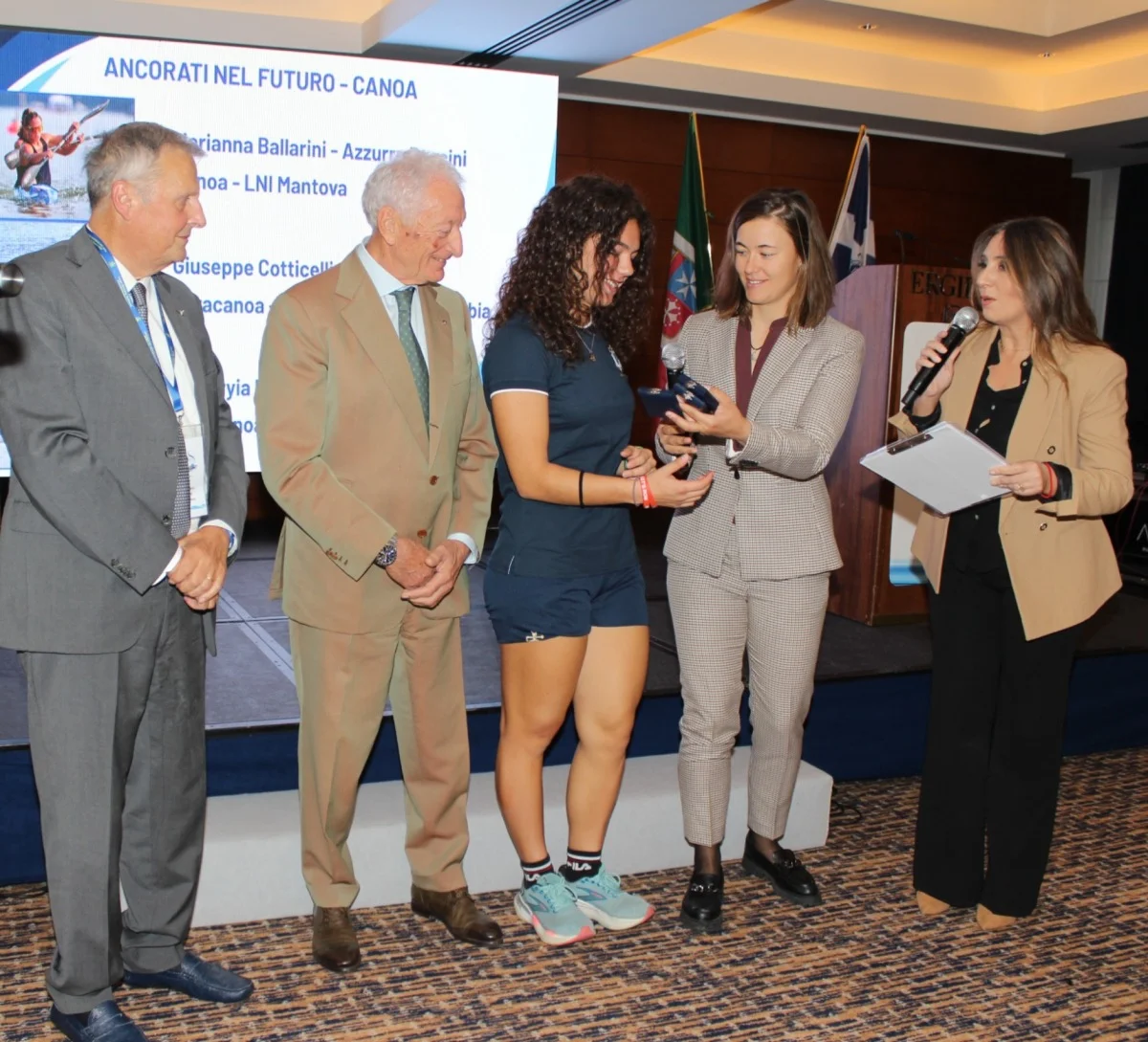Premiazione 'Ancorati nel futuro - Canoa': una giovane atleta riceve un premio da dirigenti e ospiti, con la presentatrice al microfono.