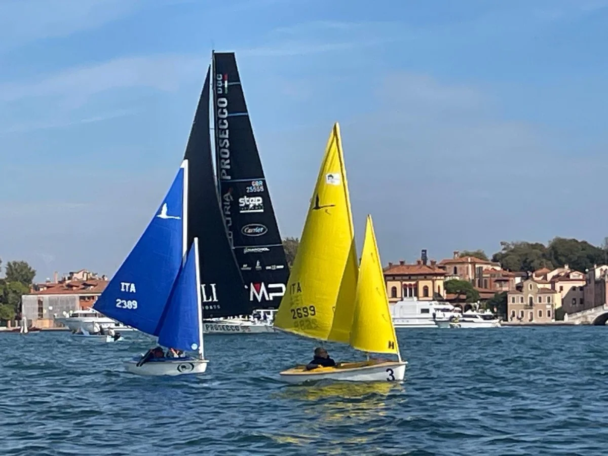 Barche a vela blu e gialle in primo piano e maxi yacht nero con sponsor alle spalle, laguna di Venezia. Molto simile alla precedente.