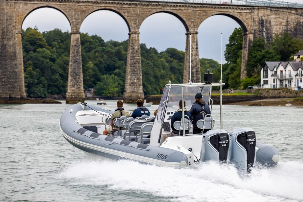 Gommon e poppa in planata con piloti a bordo, vista ravvicinata del battello e doppio fuoribordo, sfondo viadotto ad archi.