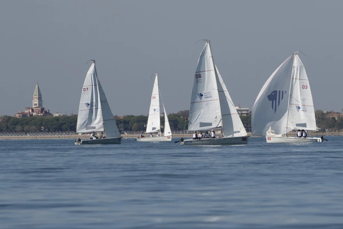Flotta di derive con vele bianche in regata in laguna, con il campanile di Venezia sullo sfondo.