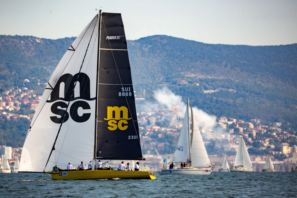Stessa barca gialla con grande vela MSC durante la regata, altre imbarcazioni e costa sullo sfondo, utile per dettagli di evento e reportage.