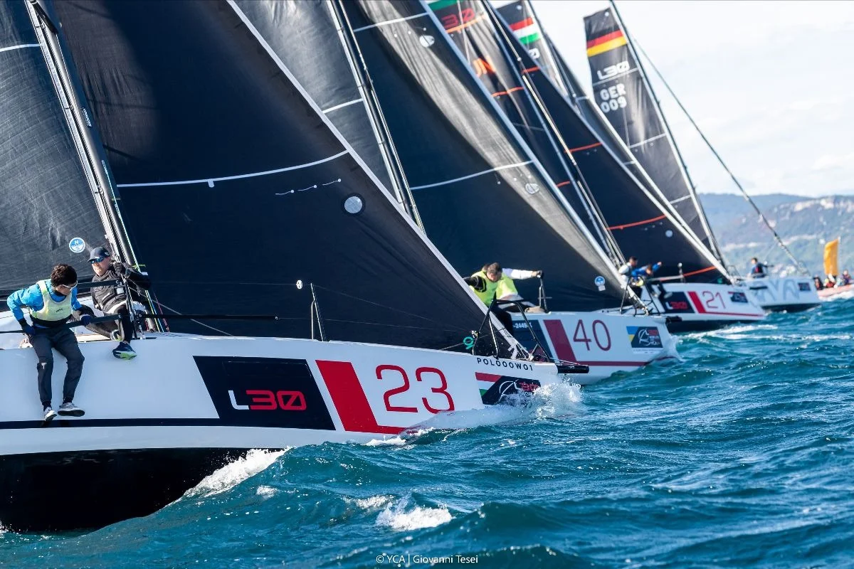 Fila di L30 in regata, prua con numeri di gara e vele nere, scena dinamica di competizione
