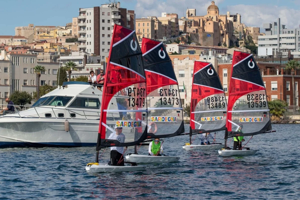 Quattro O'pen Skiff con vele rosse e nere "Sardegna" in regata davanti a un motoscafo e al centro storico di Cagliari.