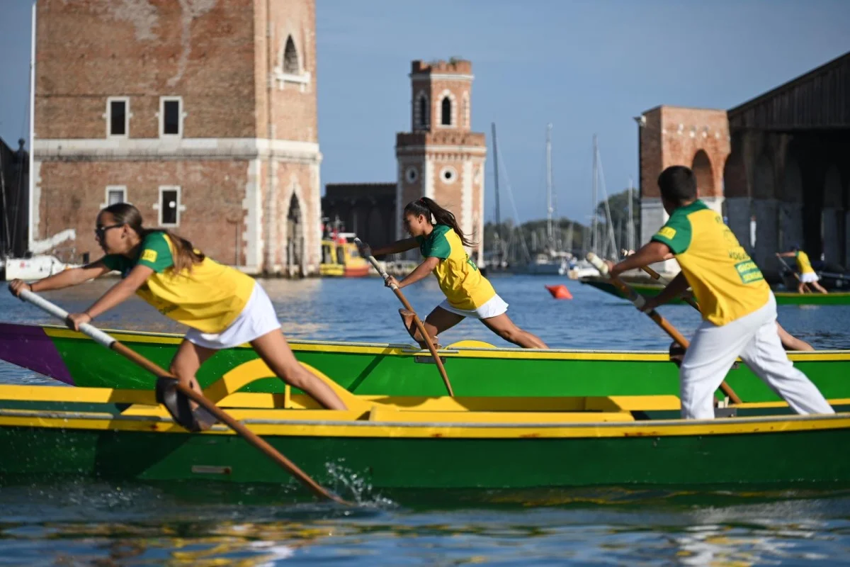 Vogatori in maglia gialloverde su imbarcazioni tradizionali davanti alle torri dell'Arsenale di Venezia.