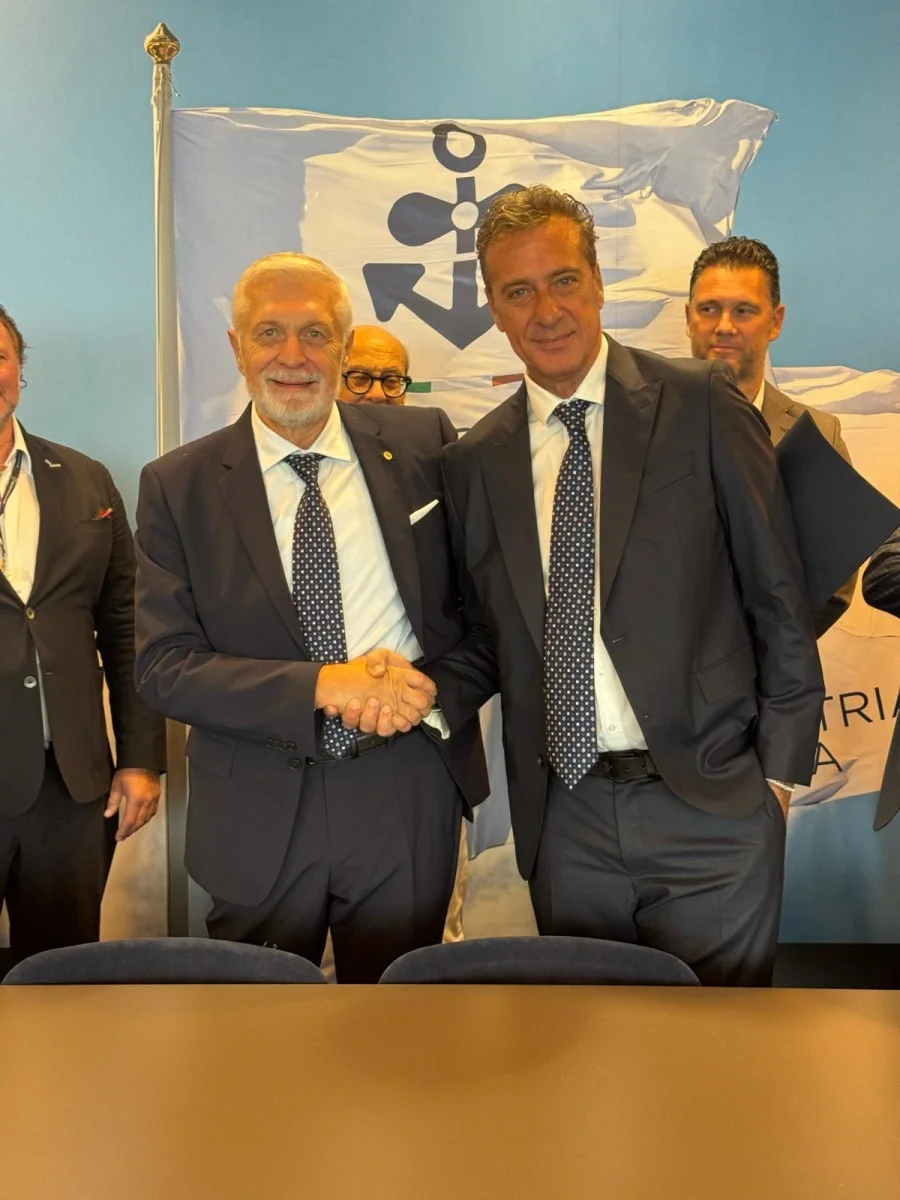 Stretta di mano tra due dirigenti davanti alla bandiera; inquadratura verticale, dettaglio di accordo o firma.