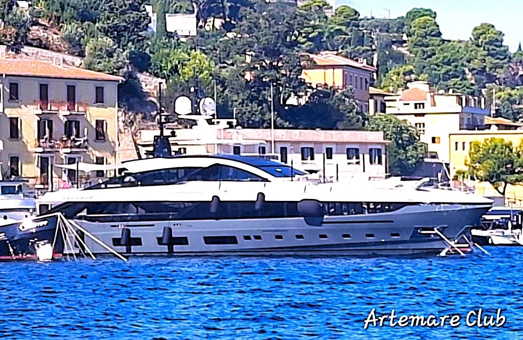 Yacht dalle linee moderne ormeggiato davanti a un borgo costiero con case colorate e vegetazione; mare blu intenso, watermark.