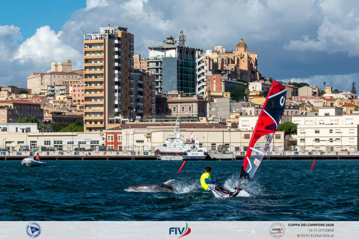 Surfer con vela rossa in primo piano, scia d&rsquo;acqua e skyline cittadino sullo sfondo