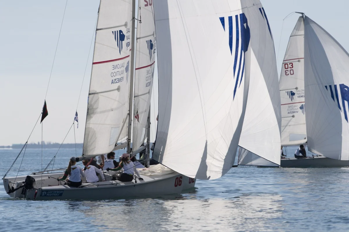 Primo piano di barche a vela del Salone Nautico di Venezia in regata, con equipaggio a bordo e vele con logo blu.