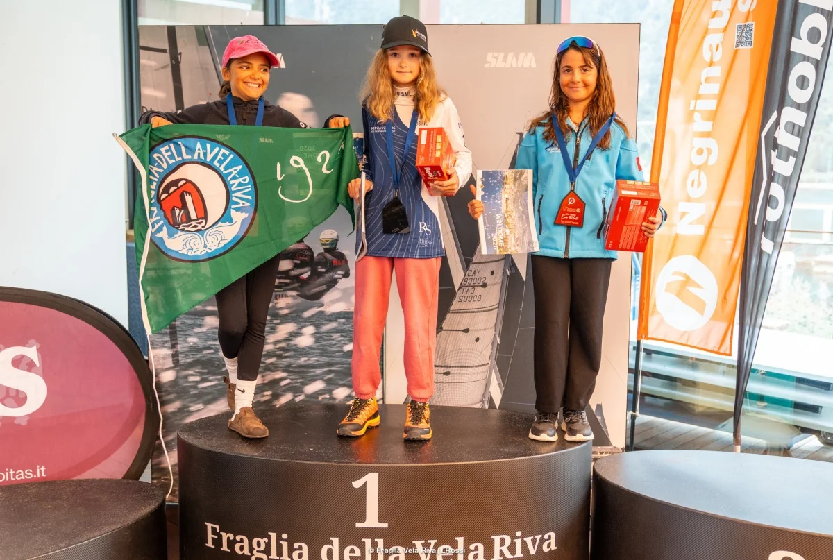 Podio di tre ragazze con medaglie, bandiera verde della Fraglia e premi, con banner sponsor.
