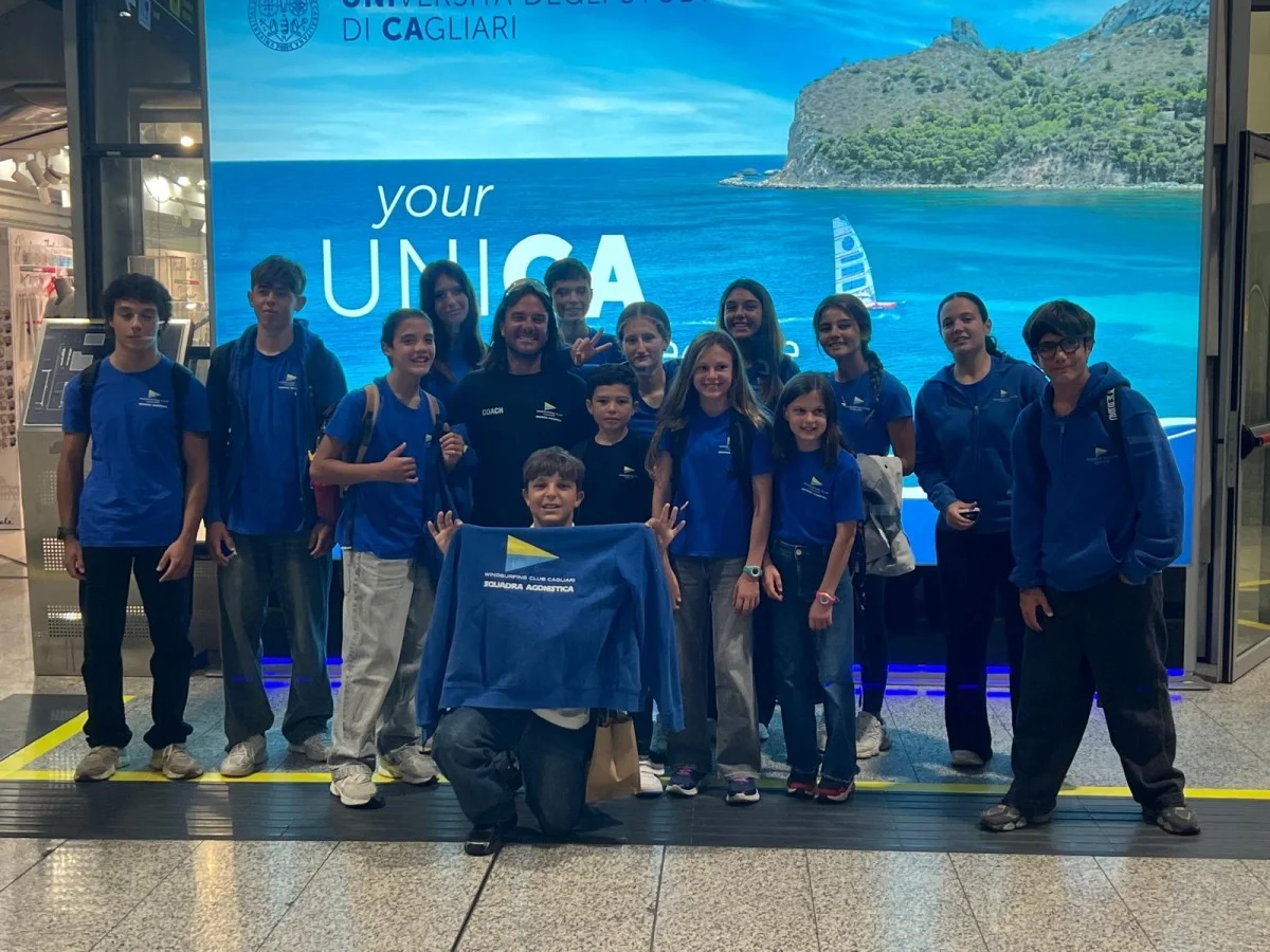 Foto di gruppo di giovani velisti in maglia blu con il coach, in aeroporto davanti a un pannello dell'Università di Cagliari.