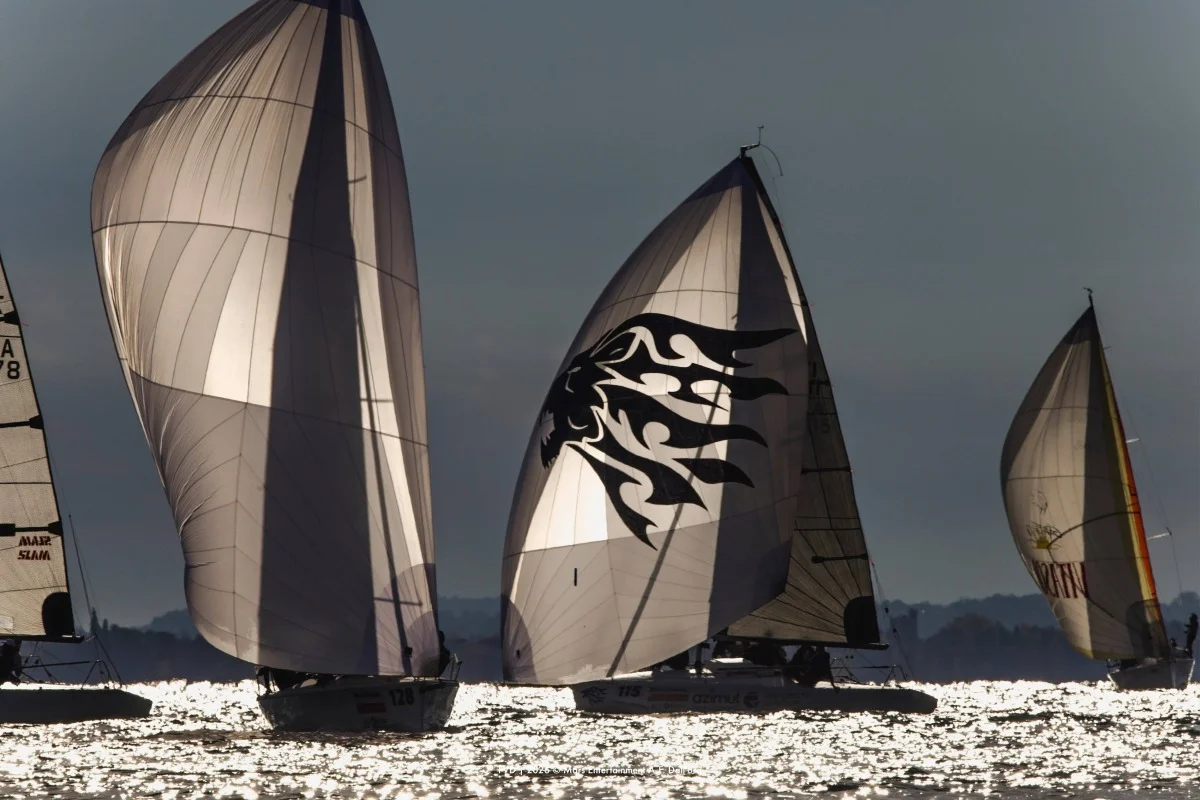 Spinnaker in controluce su barche a vela con acqua scintillante; scatto suggestivo e artistico, ma inquadratura stretta e con piccola firma.