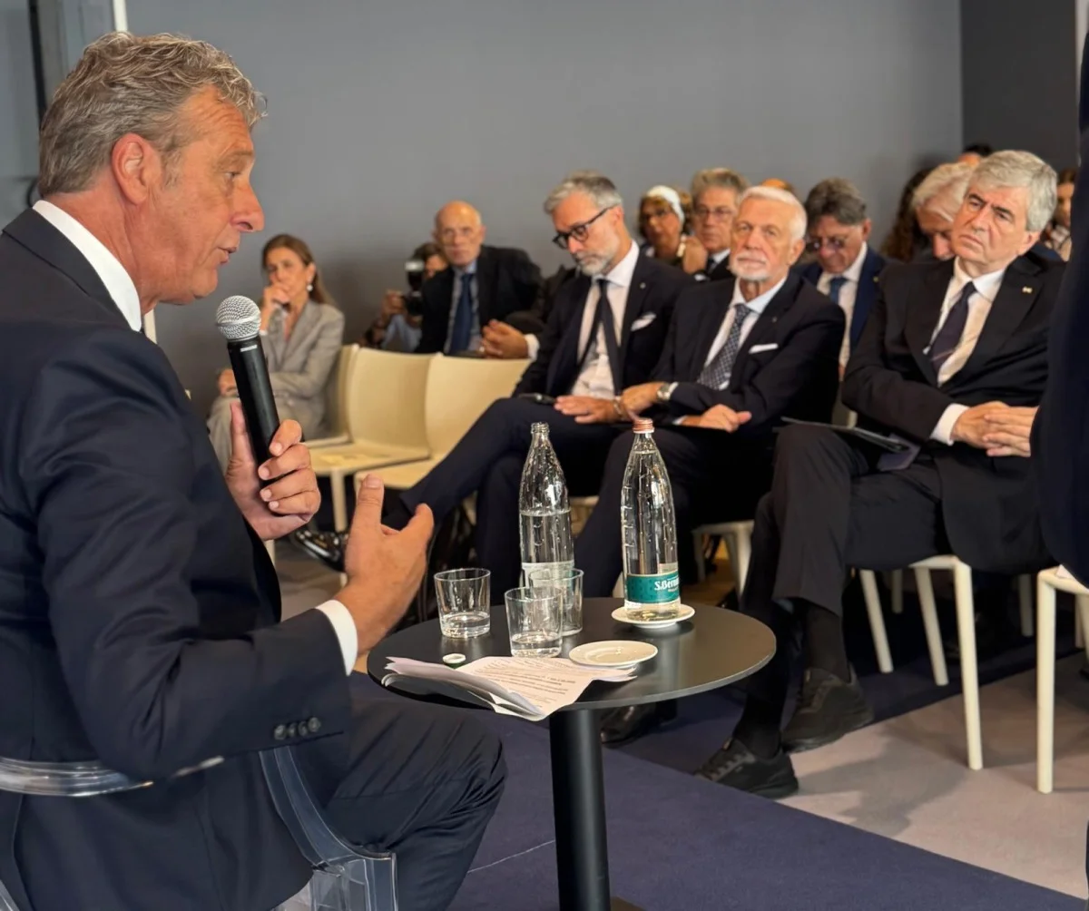 Relatore con microfono di profilo e pubblico attento seduto, scena di conferenza o tavola rotonda.