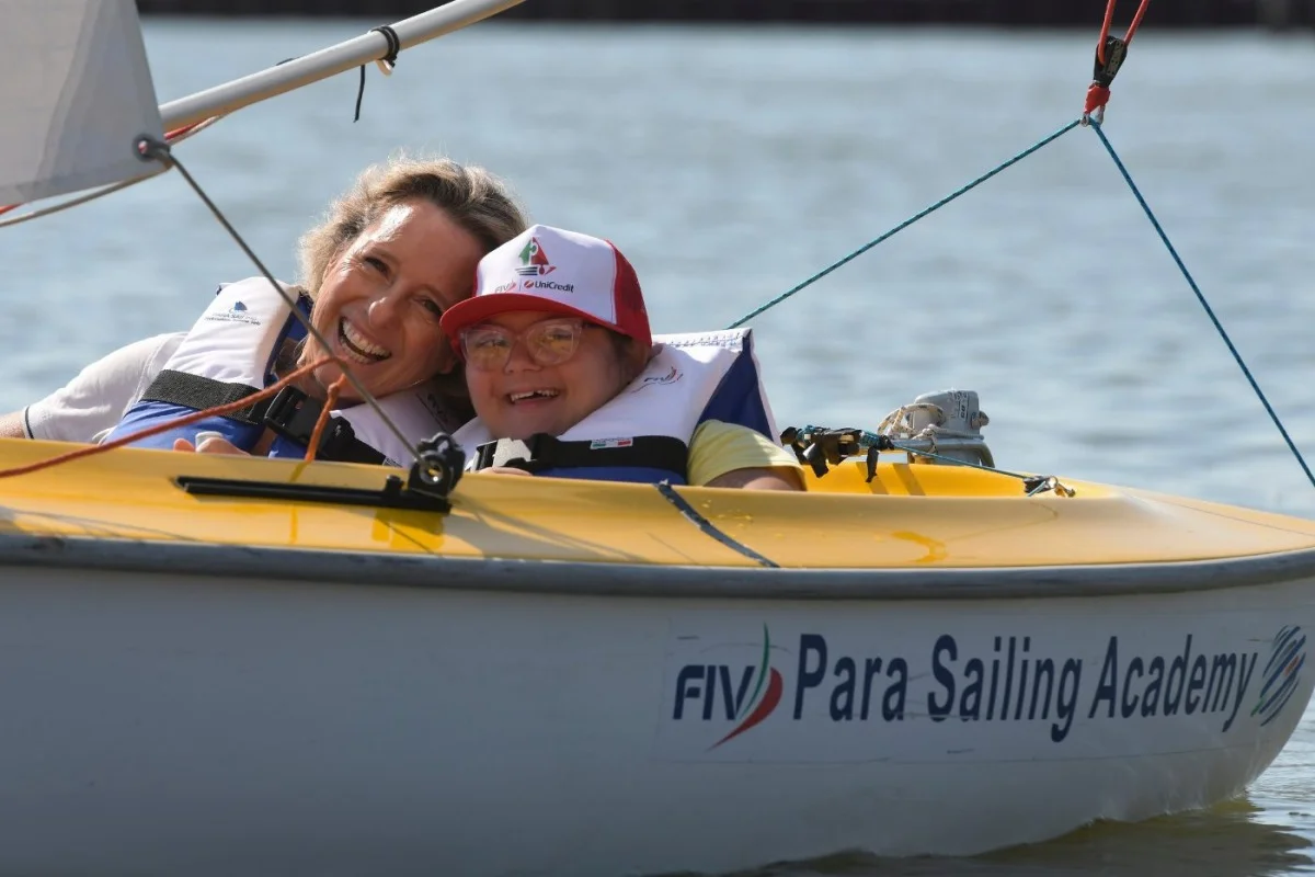 Due persone sorridenti a bordo di un piccolo dinghy giallo con scritta 'Para Sailing Academy'.