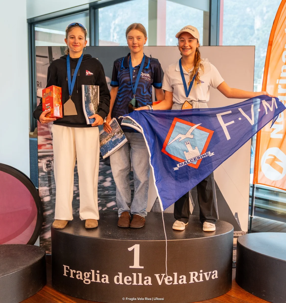 Podio di tre ragazze con medaglie, premi e guidone FVM Malcesine; formato quasi quadrato/verticale.