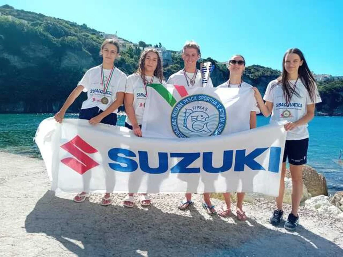 Cinque giovani atleti FIPSAS con medaglie reggono uno striscione Suzuki su una spiaggia, mare e costa sullo sfondo.