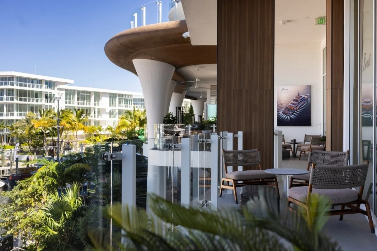 Terrazza di un edificio moderno con tavolini e sedie, palme, e un quadro di uno yacht Benetti alla parete. Probabile sede o showroom in Florida.
