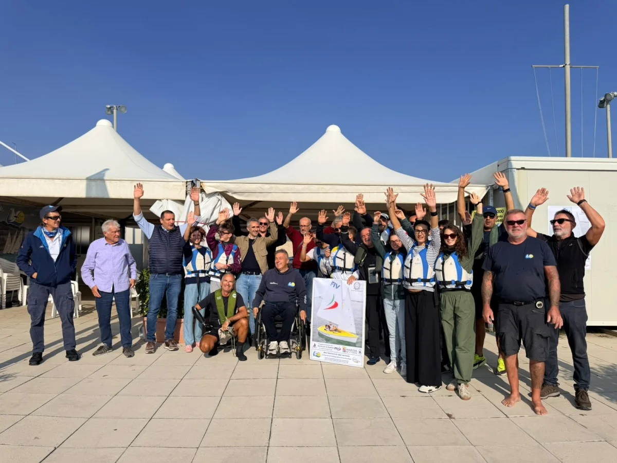 Foto di gruppo di partecipanti, istruttori e volontari con giubbotti salvagente e braccia alzate davanti ai gazebo bianchi, con una persona in carrozzina.