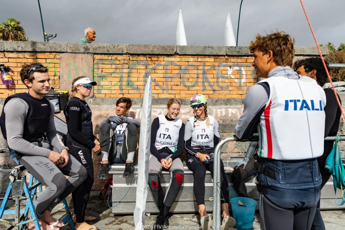 Gruppo di velisti della squadra italiana in muta e gilet ITALIA, seduti e in piedi a chiacchierare vicino a un muro con graffiti.