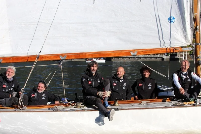 Ritratto di gruppo dell'equipaggio sorridente seduto a bordo della barca d'epoca con giacche nere.