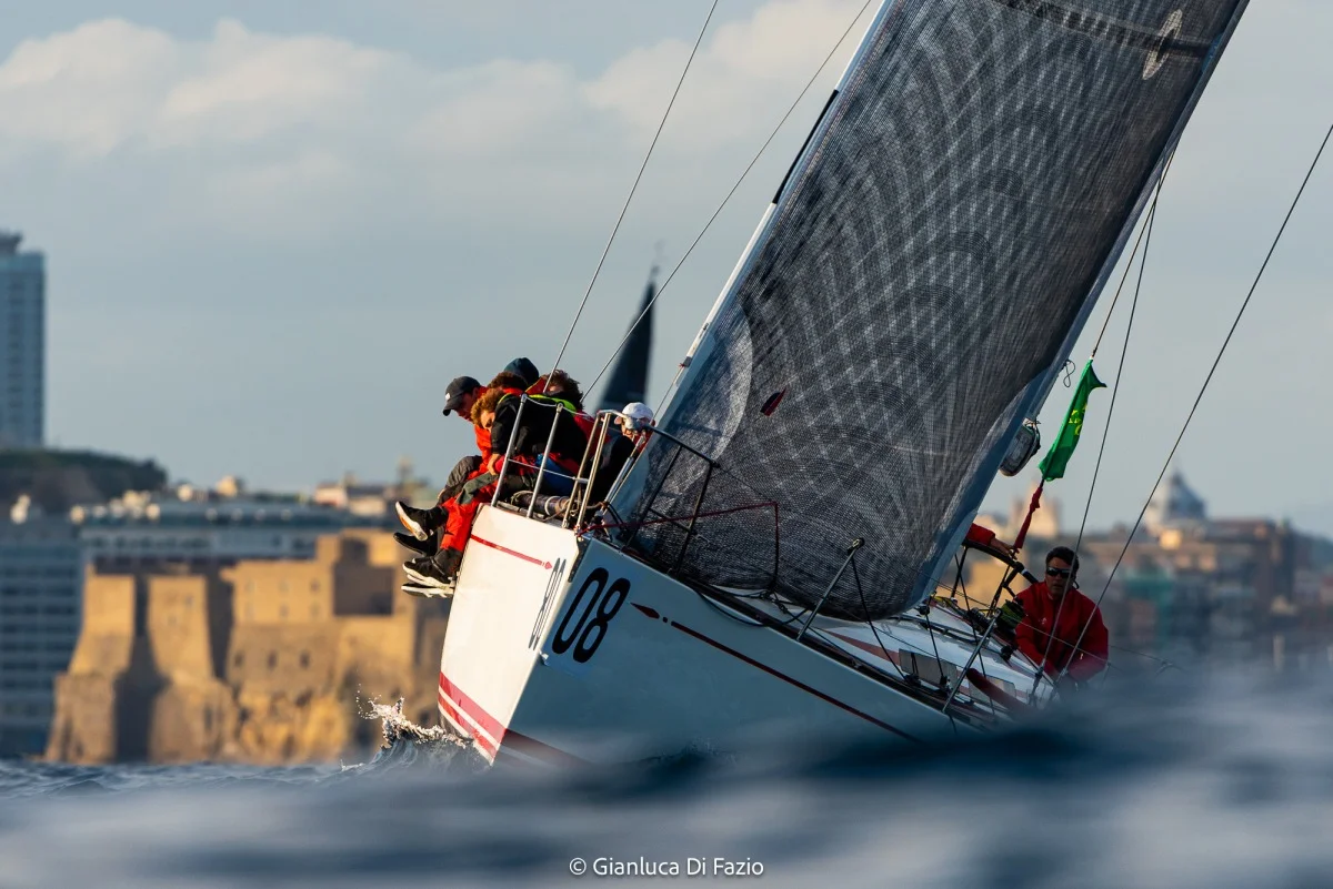 Vela da regata inclinata con equipaggio a bordo e skyline urbano sullo sfondo, prospettiva bassa sull'acqua; buona per dettagli di gara.