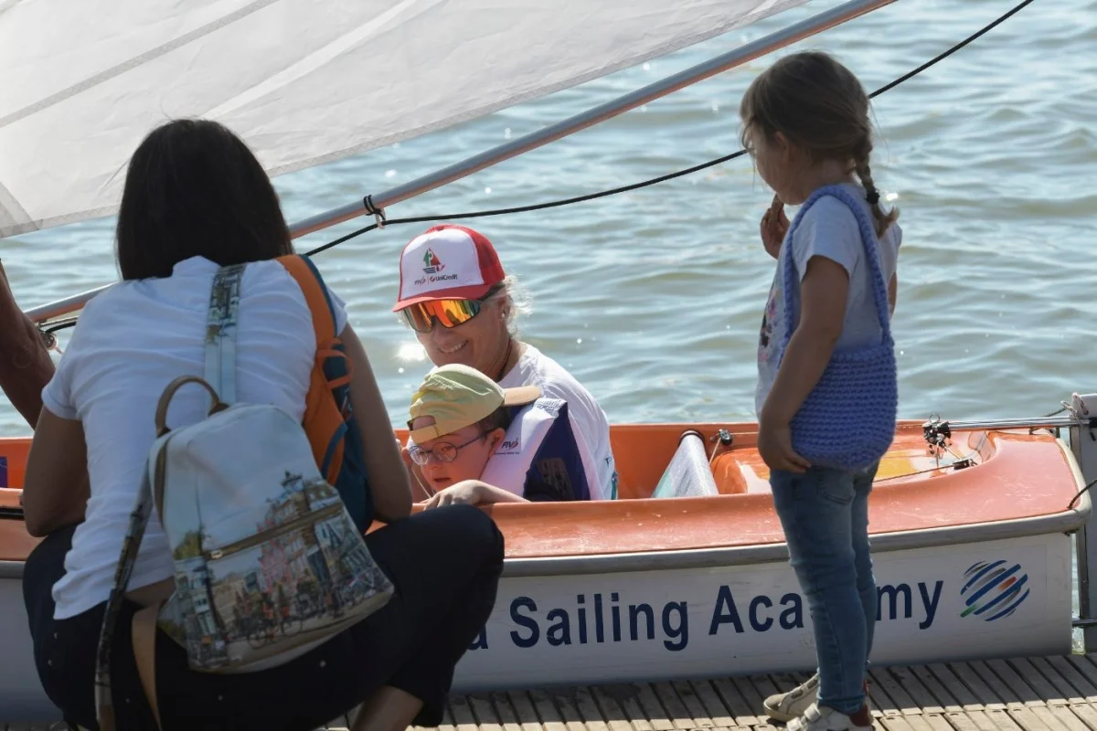 Scena al pontile: istruttrice e bambino su un dinghy con osservatori, atmosfera di attivit&agrave; didattica in acqua.