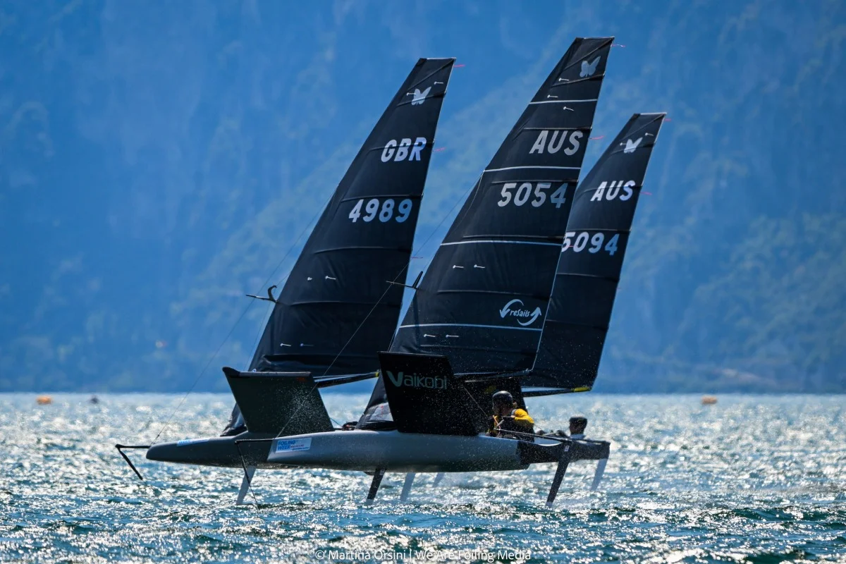 Due catamarani foiling in regata con scafo e vele nere su sfondo montano e acqua luccicante.