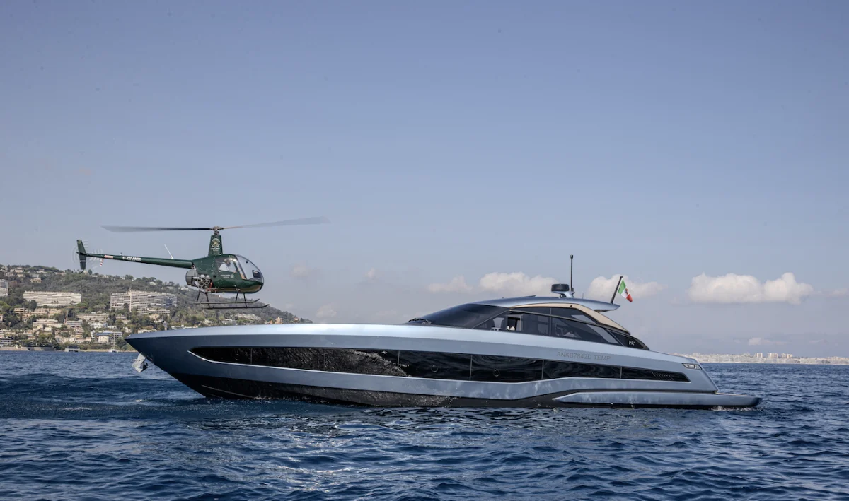 Yacht in profilo laterale sul mare con elicottero verde in volo accanto e costa sullo sfondo.