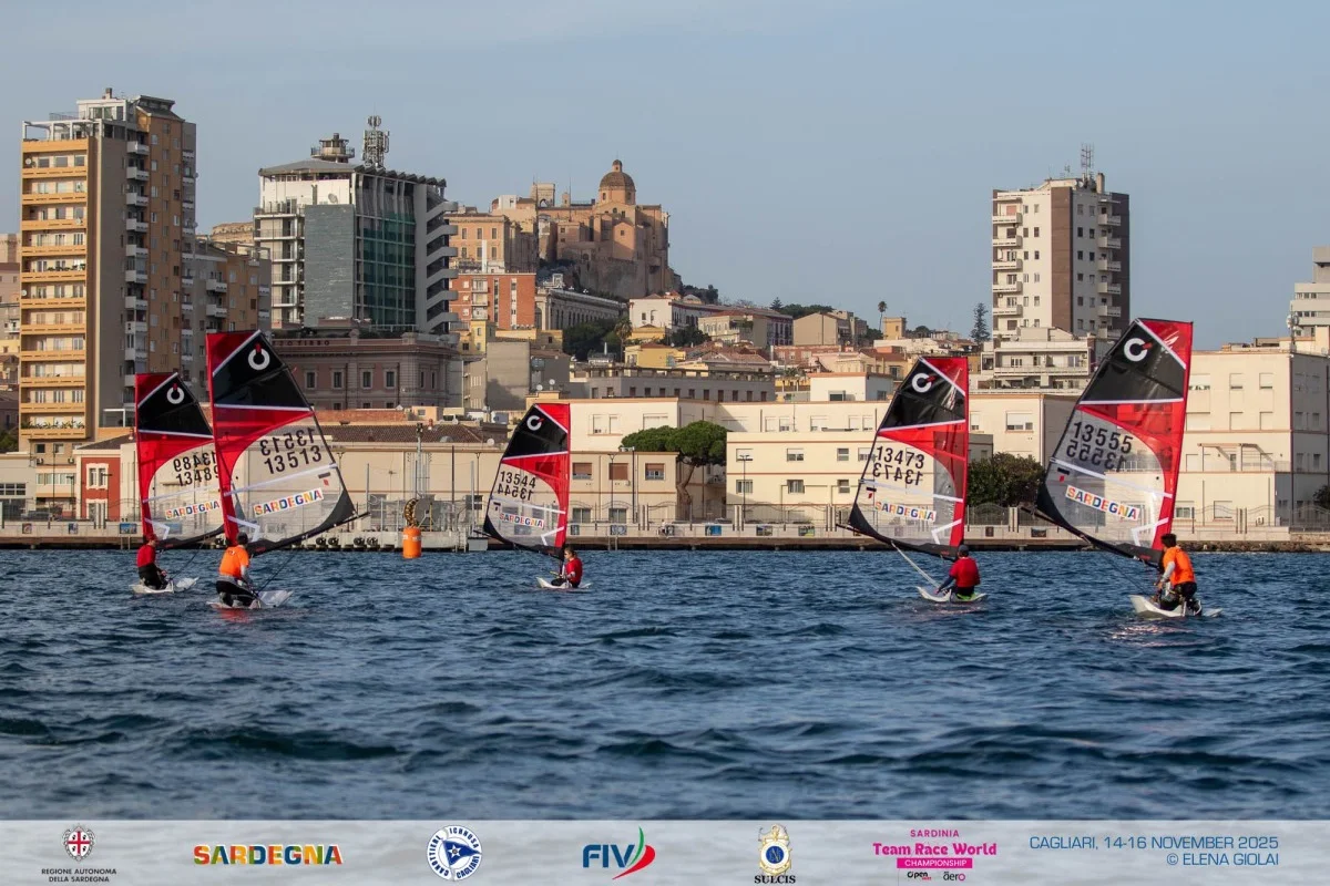 Cinque windsurfer con vele rosso-nere in navigazione davanti ai palazzi di Cagliari durante il Team Race World Championship.