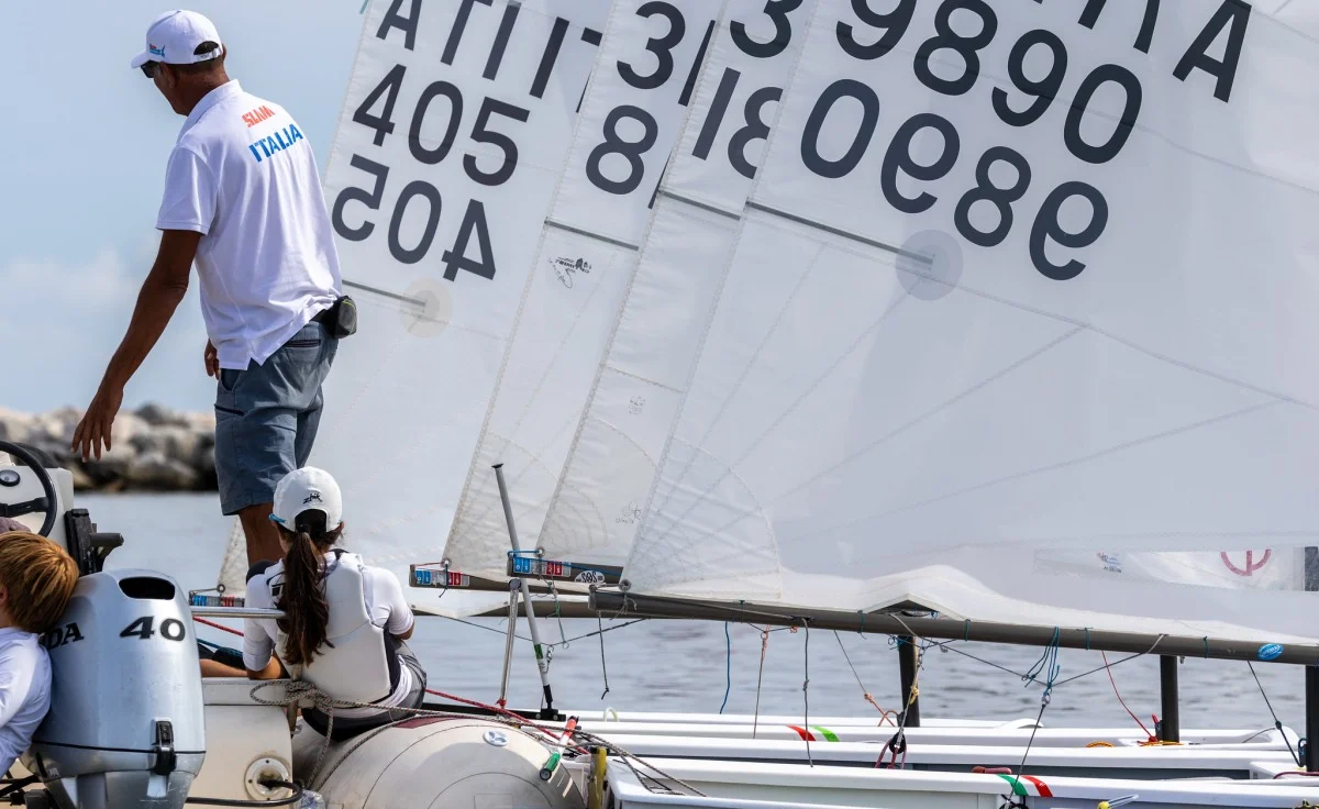 Scafo e vele di più derive con numeri, equipaggio e gommone di supporto durante la preparazione alla partenza