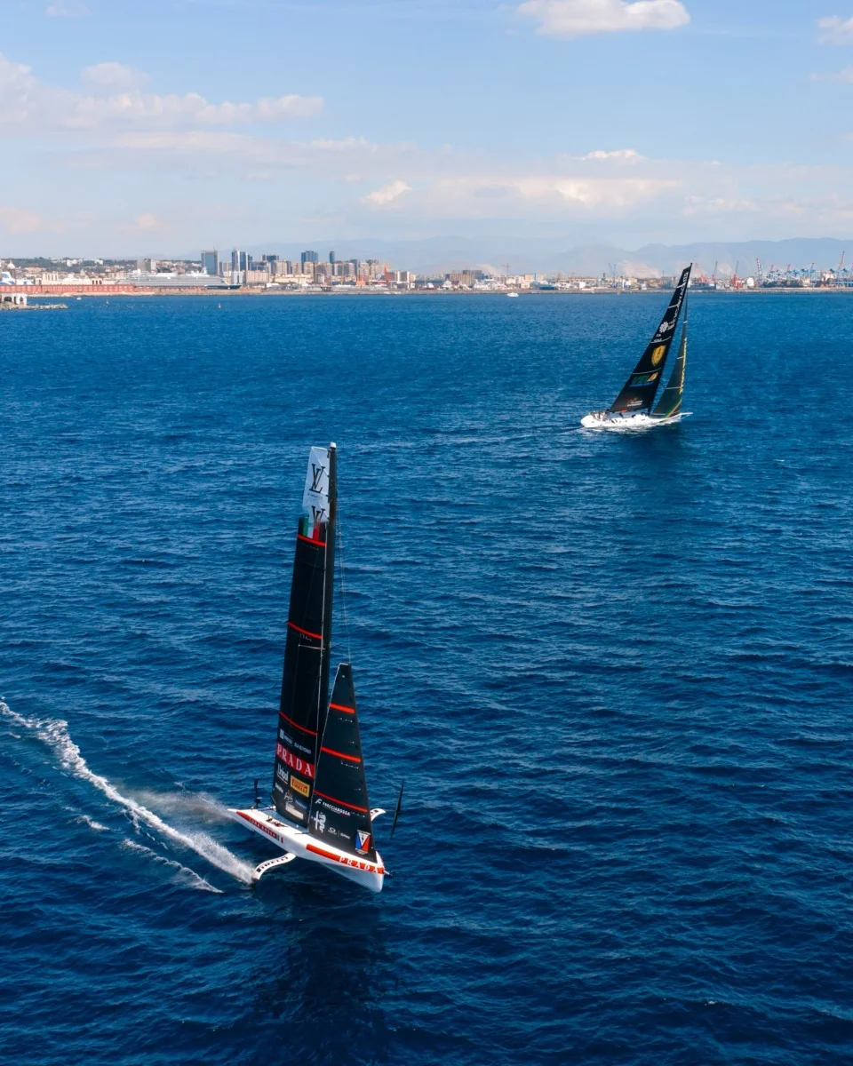 Catamarano in navigazione con città sullo sfondo; verticale ma utile per dettagli della barca