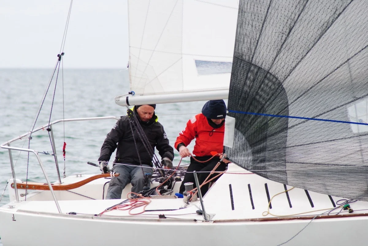 Dettaglio di due membri dell'equipaggio al lavoro a bordo, con cime e spinnaker scuro in primo piano.