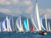 Flotta di barche a vela d'epoca con spinnaker colorati in regata davanti a una costa collinare, sotto un cielo con nuvole