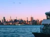 Skyline urbano al tramonto visto dall'acqua, con un traghetto in primo piano a destra.